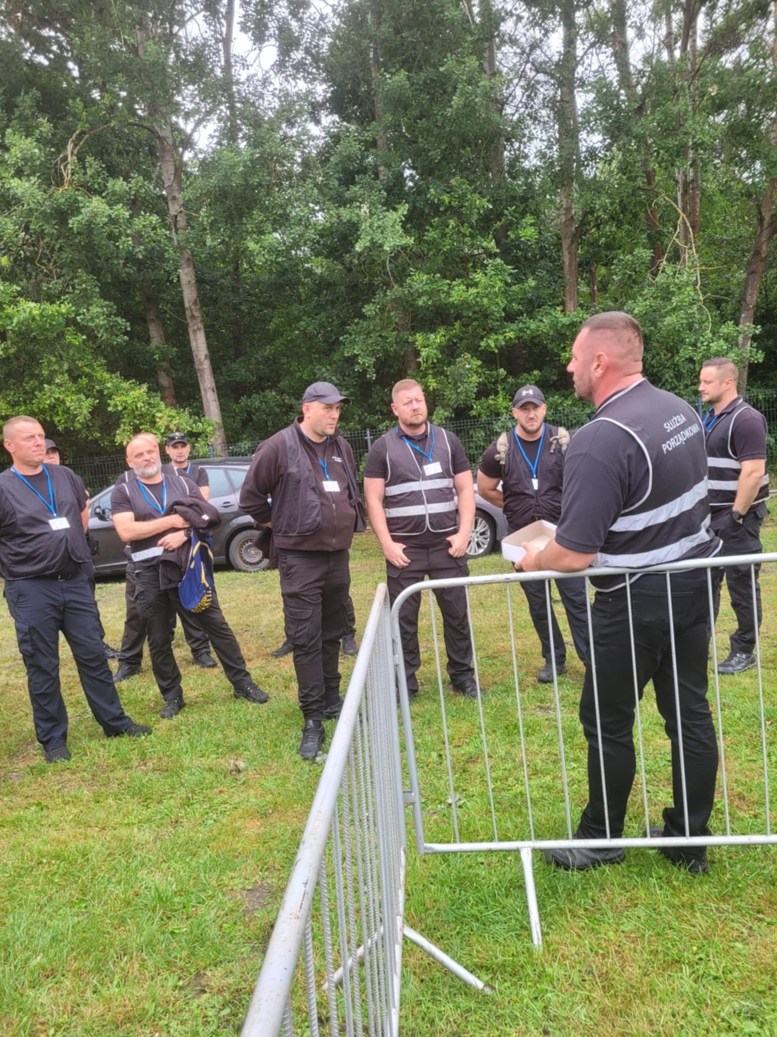 Grupa pracowników ochrony w czarnych uniformach i odblaskowych kamizelkach podczas odprawy na trawniku przed terenem zielonym. Widoczny fragment metalowego ogrodzenia.