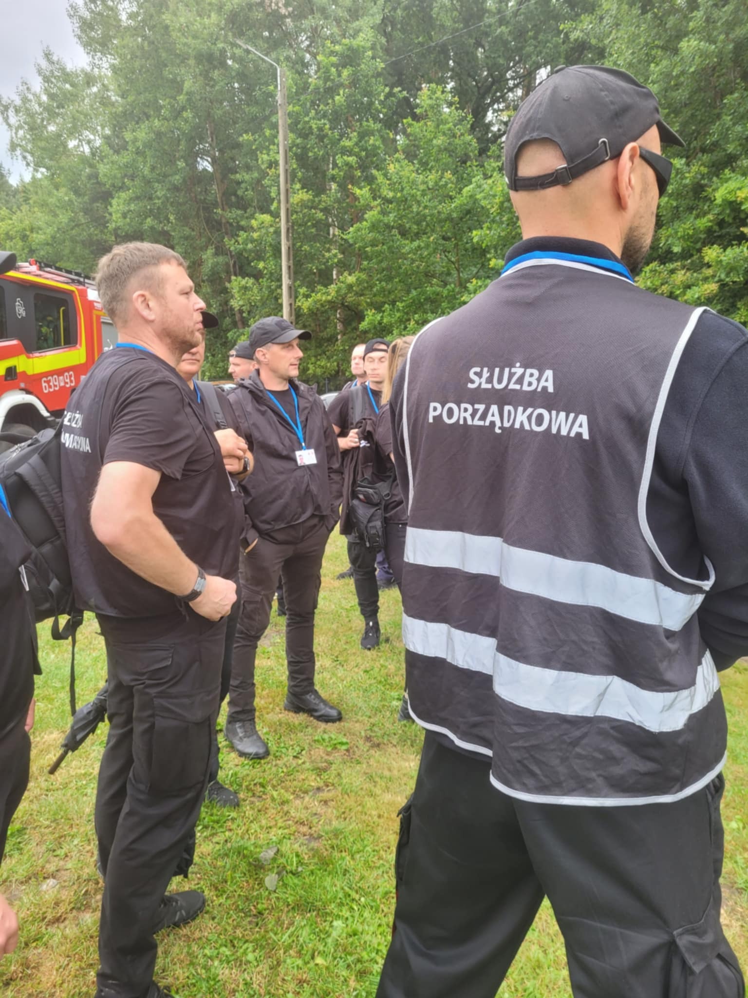 Grupa umundurowanych mężczyzn z napisem 'Służba Porządkowa' na kamizelkach, na tle zieleni i wozu strażackiego. Teren zielony, dzień.
