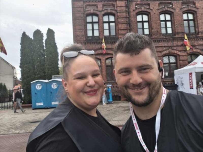 Uśmiechnięta kobieta i mężczyzna na tle ceglanego budynku z flagami i przenośnych toalet. Mężczyzna ma słuchawkę w uchu i smycz na szyi.