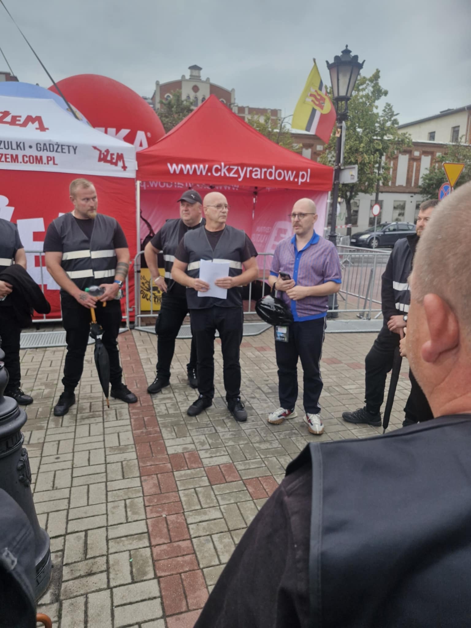 Grupa pracowników w kamizelkach odblaskowych podczas briefingu na zewnątrz, w tle widoczne namioty i flaga miejska Żyrardowa.