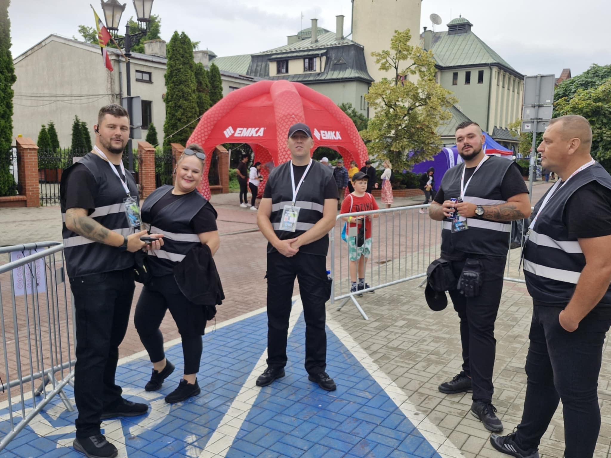 Grupa pięciu osób w strojach ochroniarzy, stojących na tle czerwonego namiotu z logo 'EMKA' i budynku z wieżą, prawdopodobnie podczas zabezpieczania imprezy plenerowej.