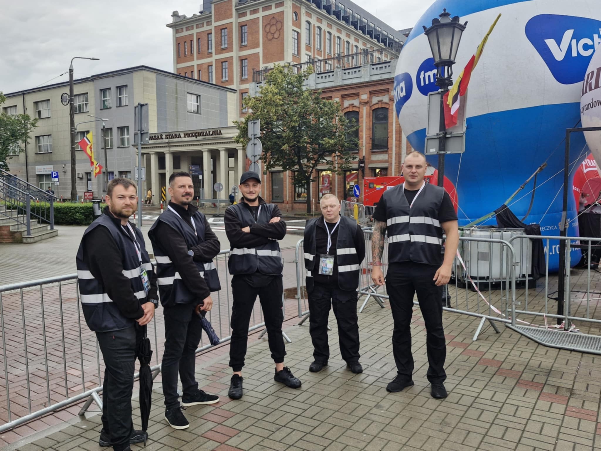 Grupa pięciu mężczyzn w czarnych uniformach i odblaskowych kamizelkach na tle miejskiej architektury i balonu reklamowego, sugerująca ochronę imprezy masowej.