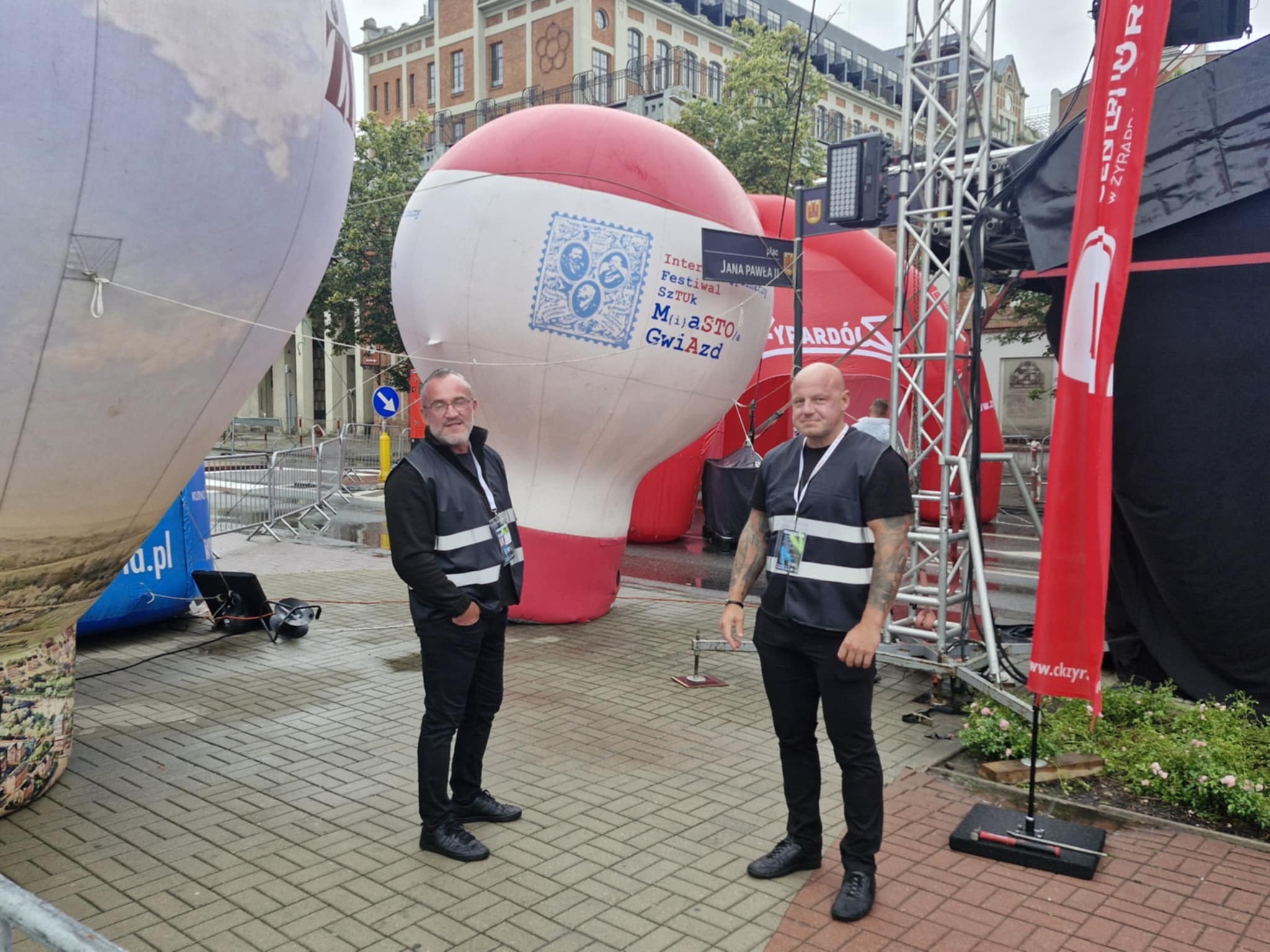 Dwóch mężczyzn w kamizelkach odblaskowych stoi na tle dekoracji festiwalowych: balony z logo i czerwony baner. Widoczne budynki w tle i bruk na placu.