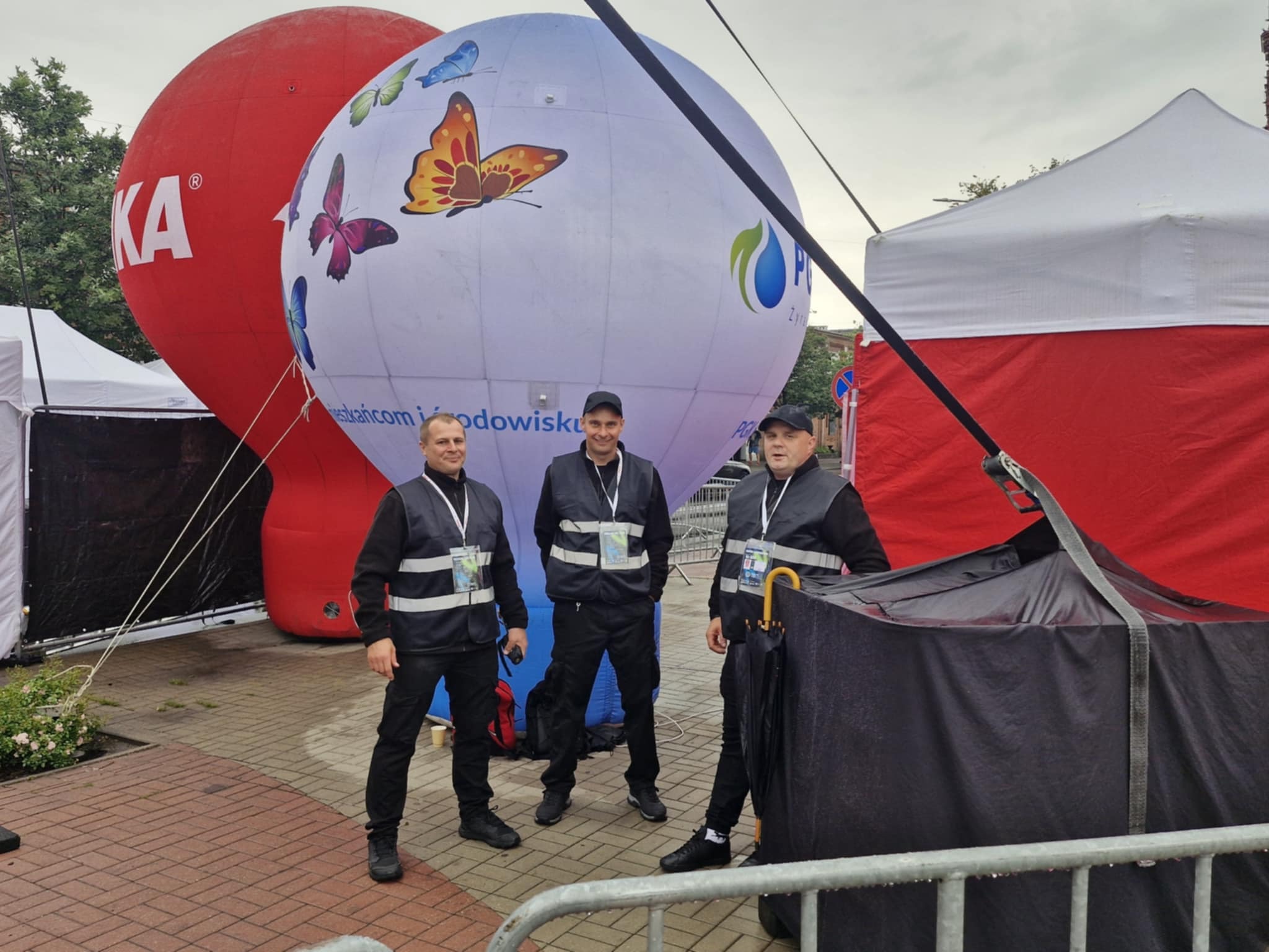 Trzech umundurowanych ochroniarzy na tle czerwonego i białego balonu z motylami, w trakcie zabezpieczania imprezy plenerowej. Widoczny fragment sceny i barierek.