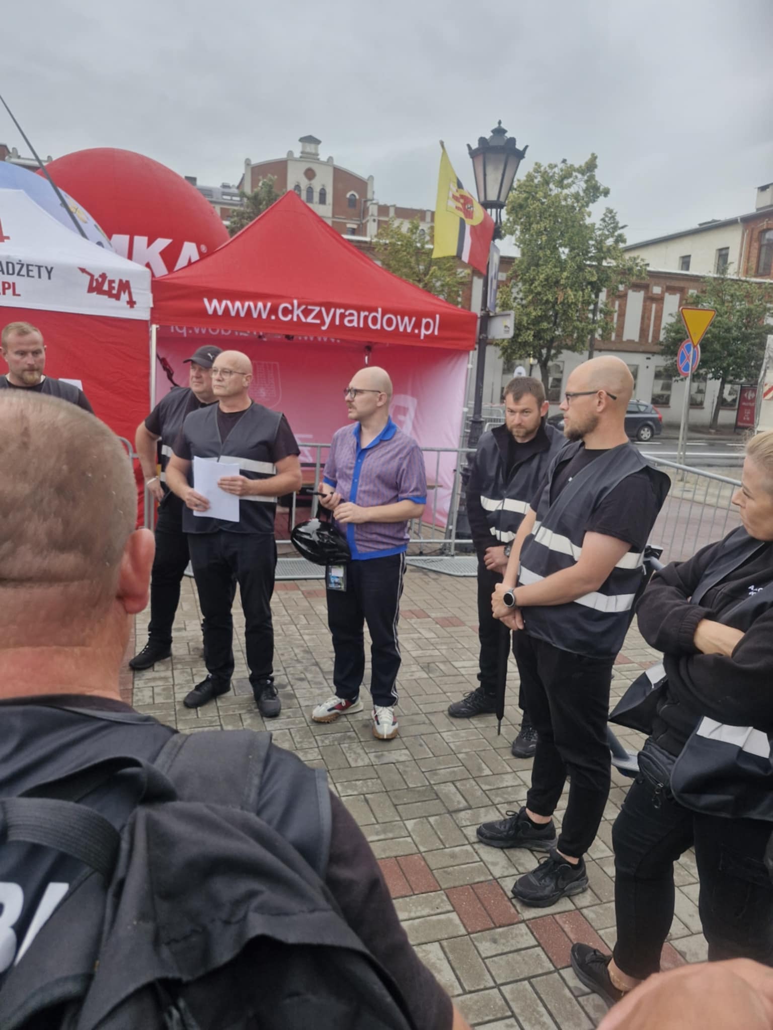 Briefing zespołu ochrony w Skierniewicach przed wydarzeniem plenerowym. Uczestnicy w kamizelkach odblaskowych i strojach roboczych. W tle czerwony namiot.