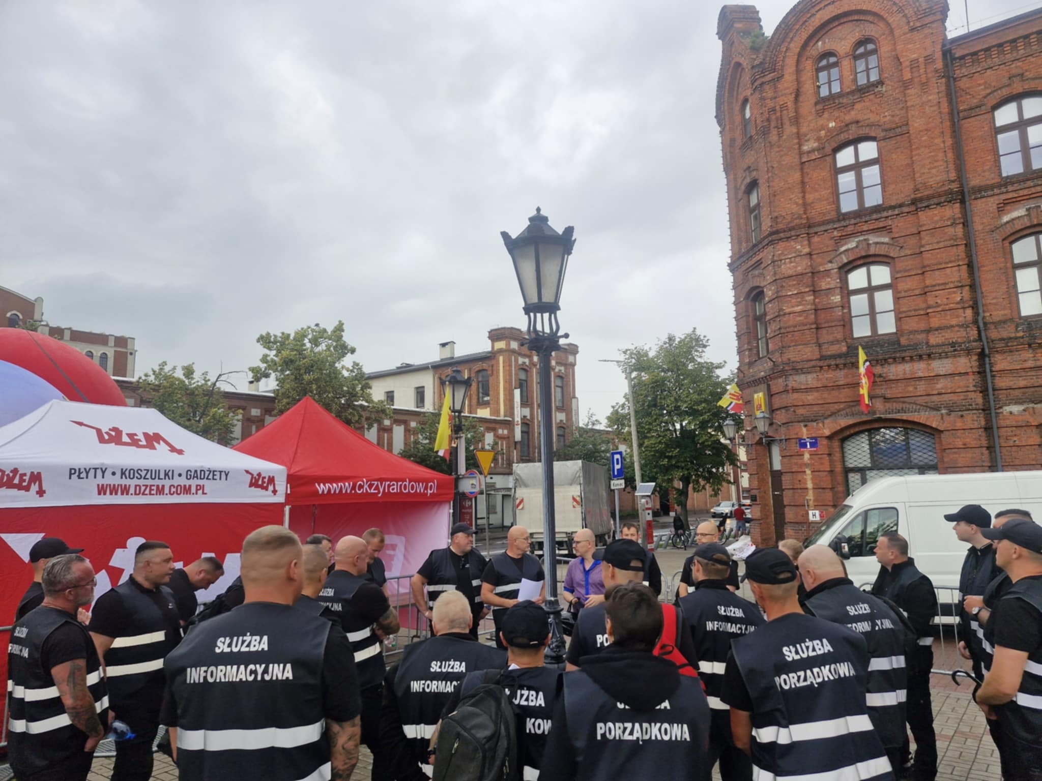 Grupa ochroniarzy w czarnych uniformach z napisem 'Służba Informacyjna' i 'Służba Porządkowa' na placu miejskim przed budynkiem z czerwonej cegły.