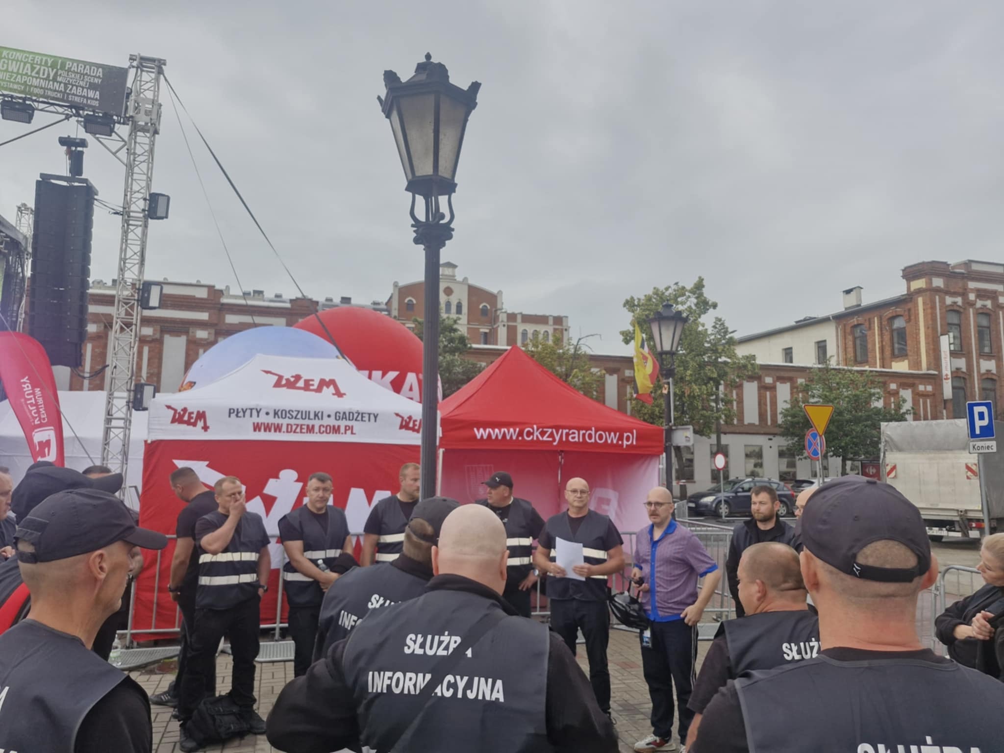 Grupa osób w kamizelkach 'Służba Informacyjna' na tle sceny i namiotów podczas wydarzenia plenerowego w Zyrardowie. Widoczna latarnia uliczna i budynki w tle.