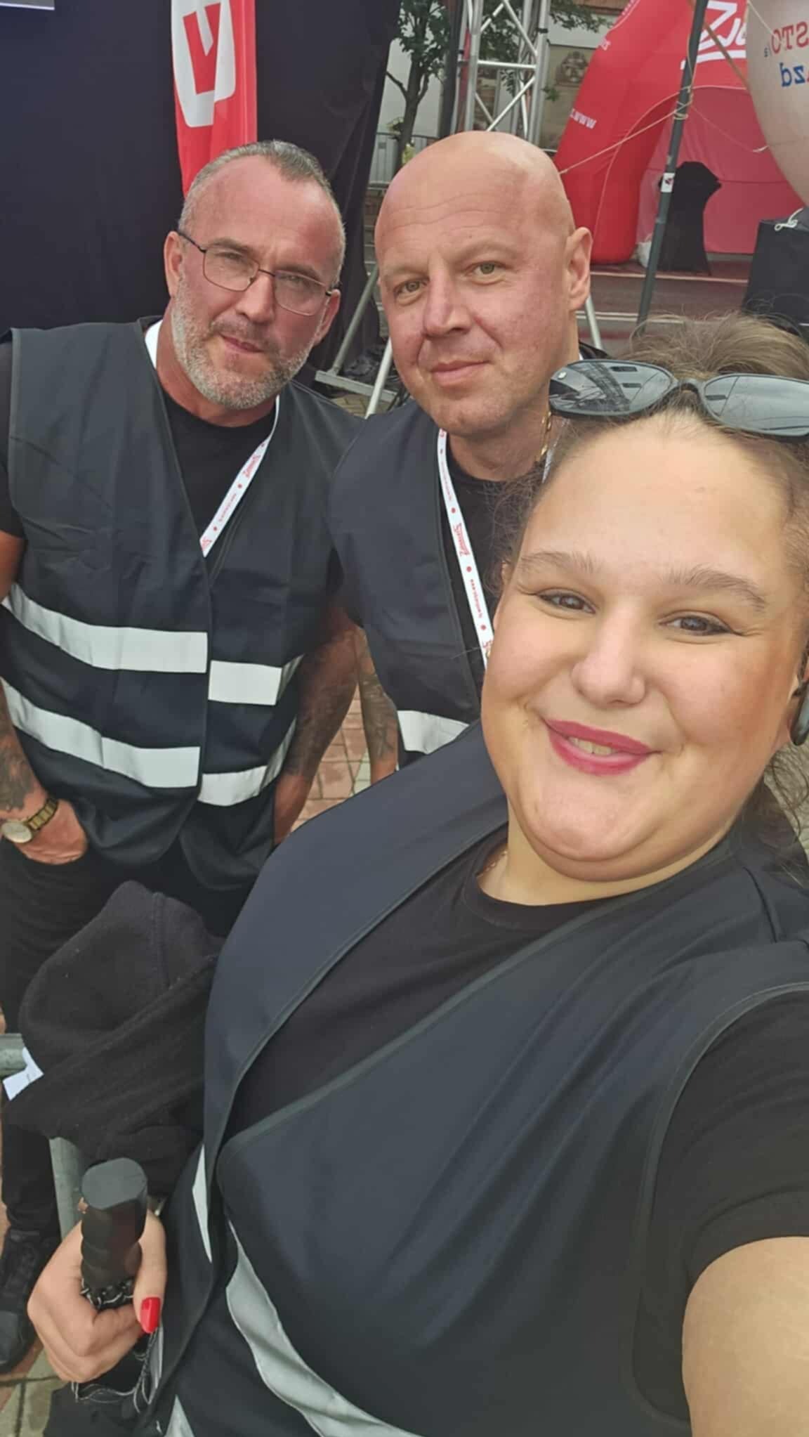 Selfie trzech osób w kamizelkach z pasami odblaskowymi na tle sceny plenerowej. Mężczyźni z brodami, kobieta z okularami na głowie. Widoczne identyfikatory.