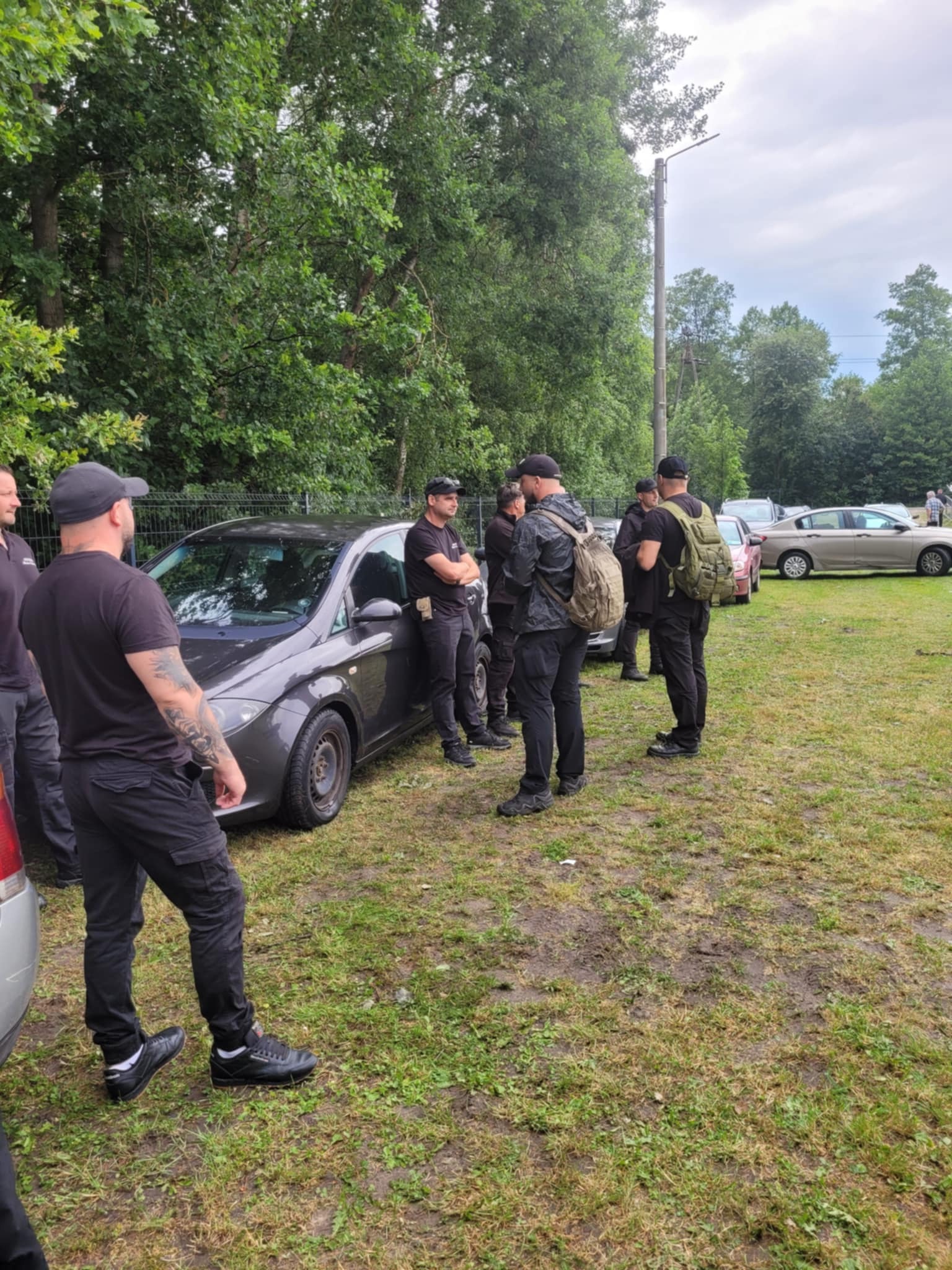 Grupa mężczyzn w czarnych ubraniach, prawdopodobnie pracowników ochrony, stoi obok szarego samochodu na trawiastym terenie w otoczeniu drzew.