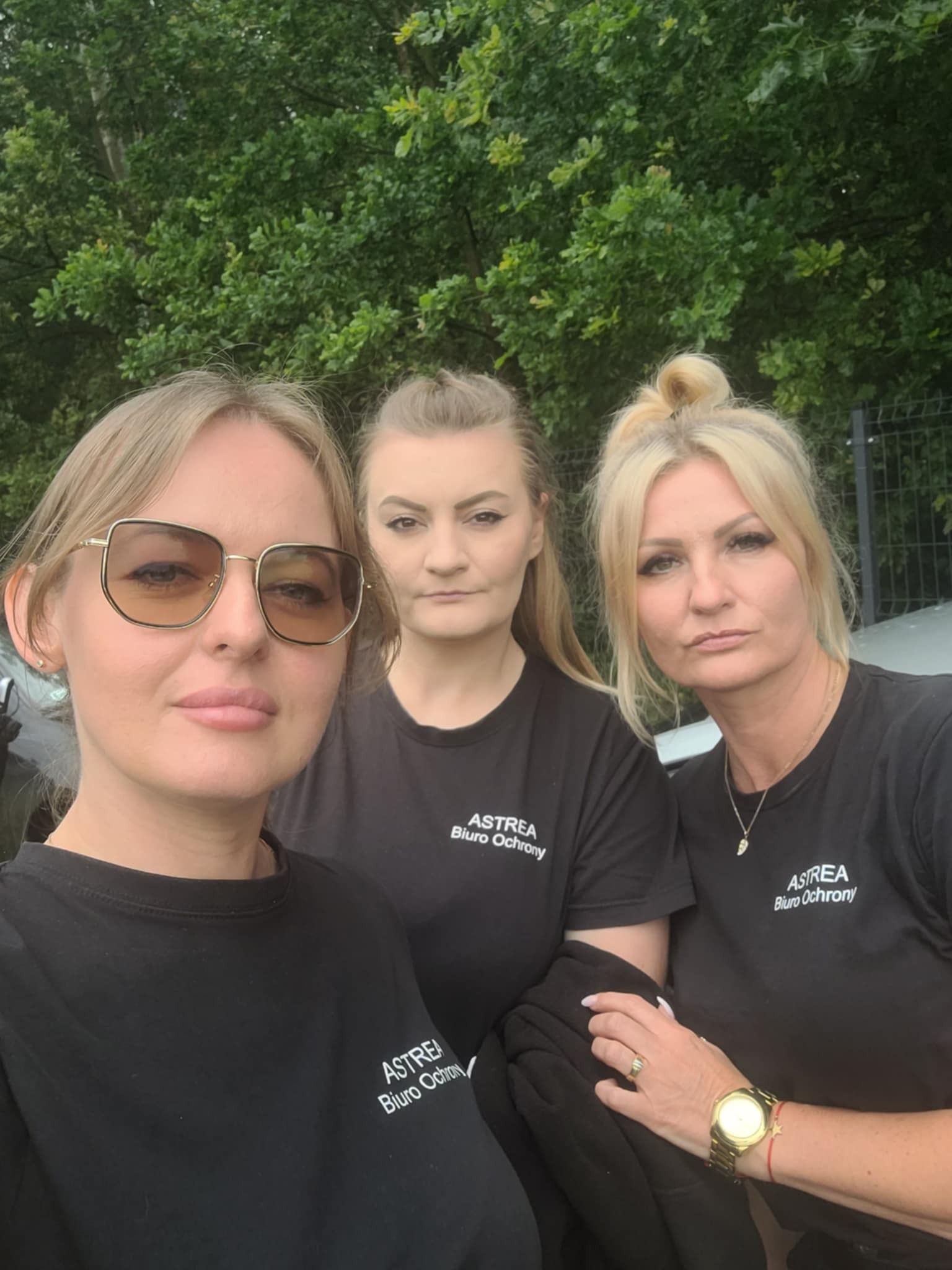 Trzy kobiety w czarnych koszulkach z logo firmy. Selfie na tle zieleni. Zegarek na ręce, okulary przeciwsłoneczne. Profesjonalny zespół.