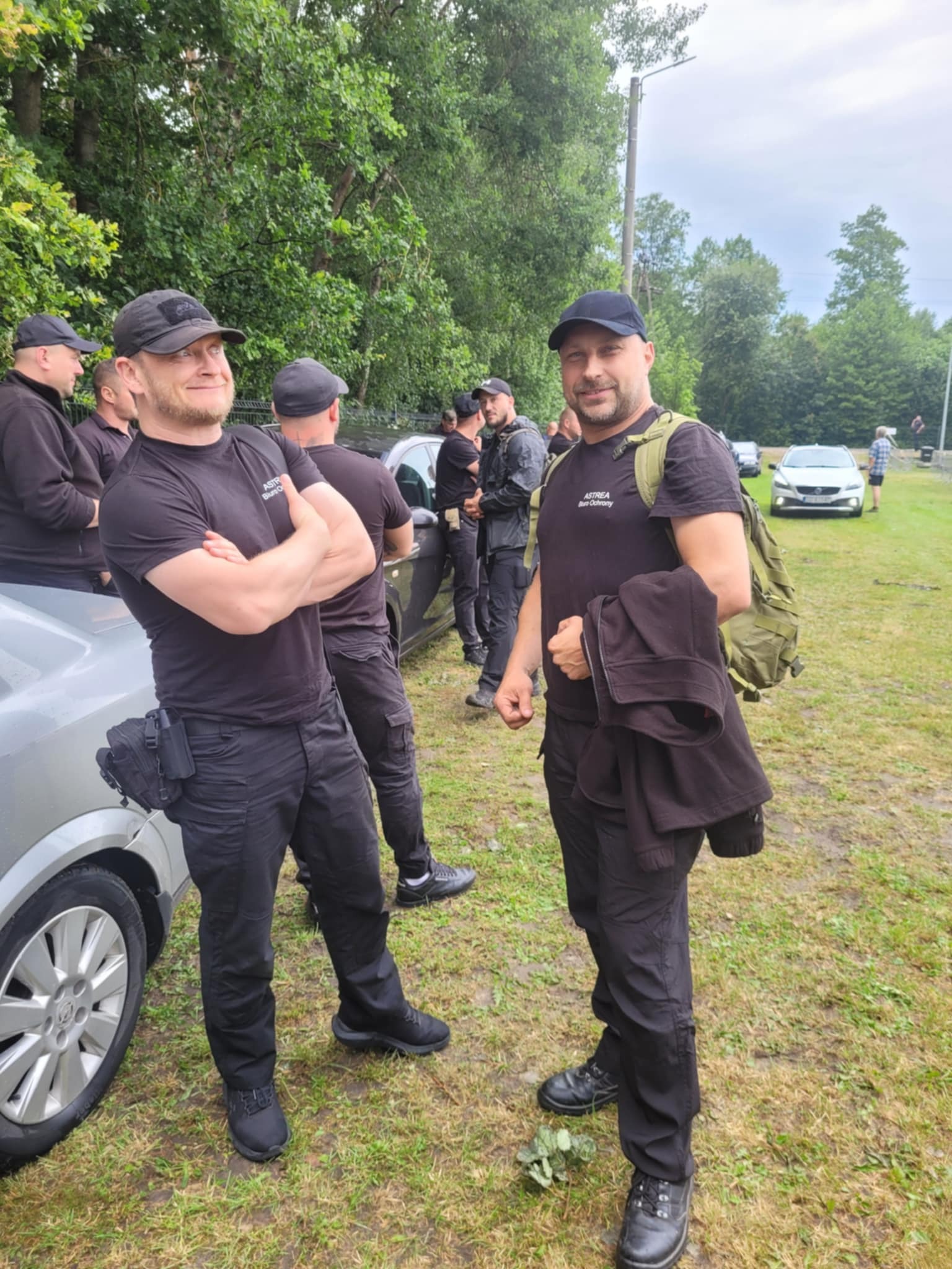 Grupa umundurowanych mężczyzn w czarnych strojach, prawdopodobnie pracowników ochrony, stojących na zewnątrz w pobliżu zaparkowanych samochodów.