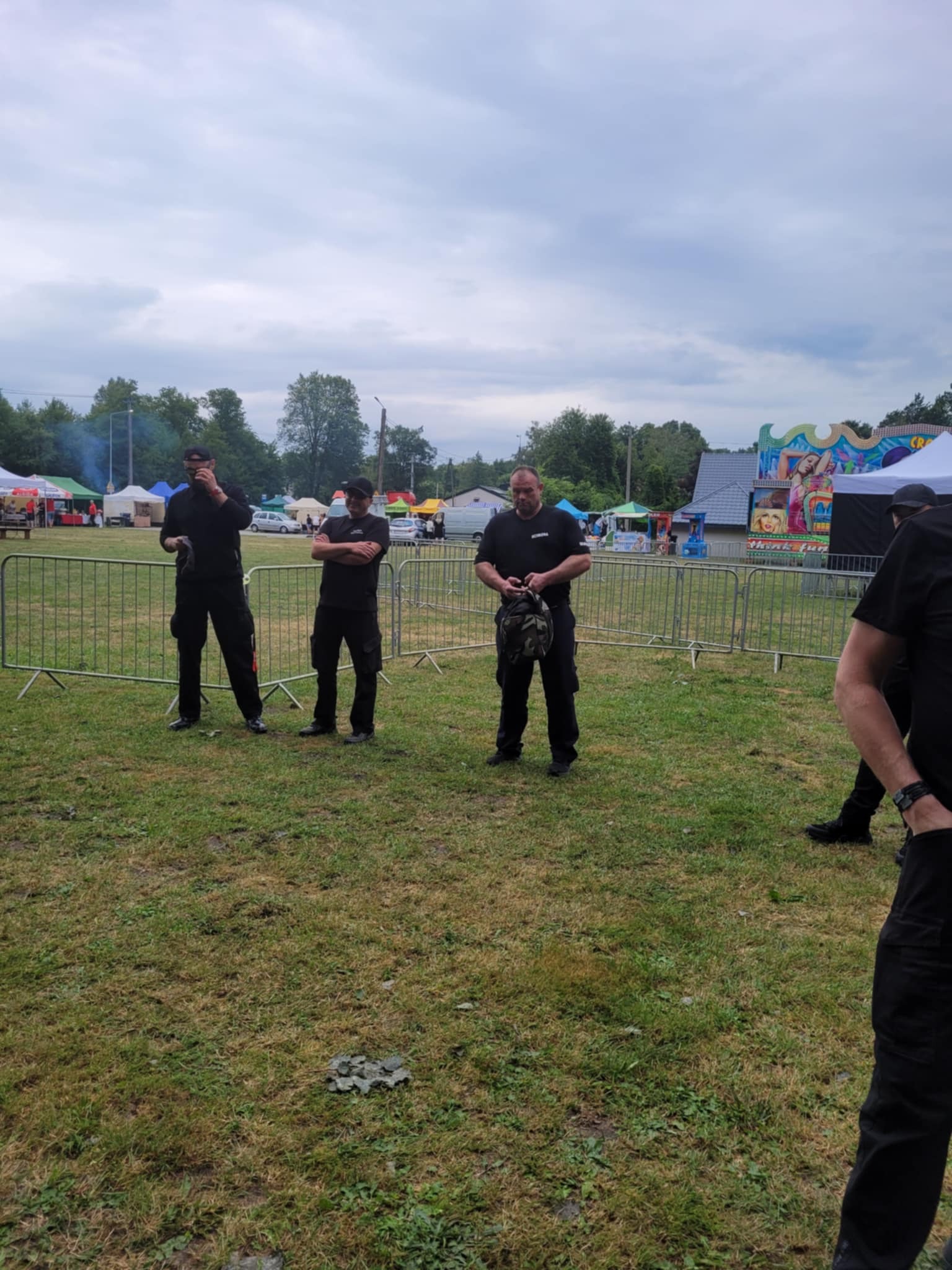 Trzech mężczyzn w czarnych uniformach stoi na trawniku podczas imprezy plenerowej, zabezpieczając teren za metalowym ogrodzeniem.