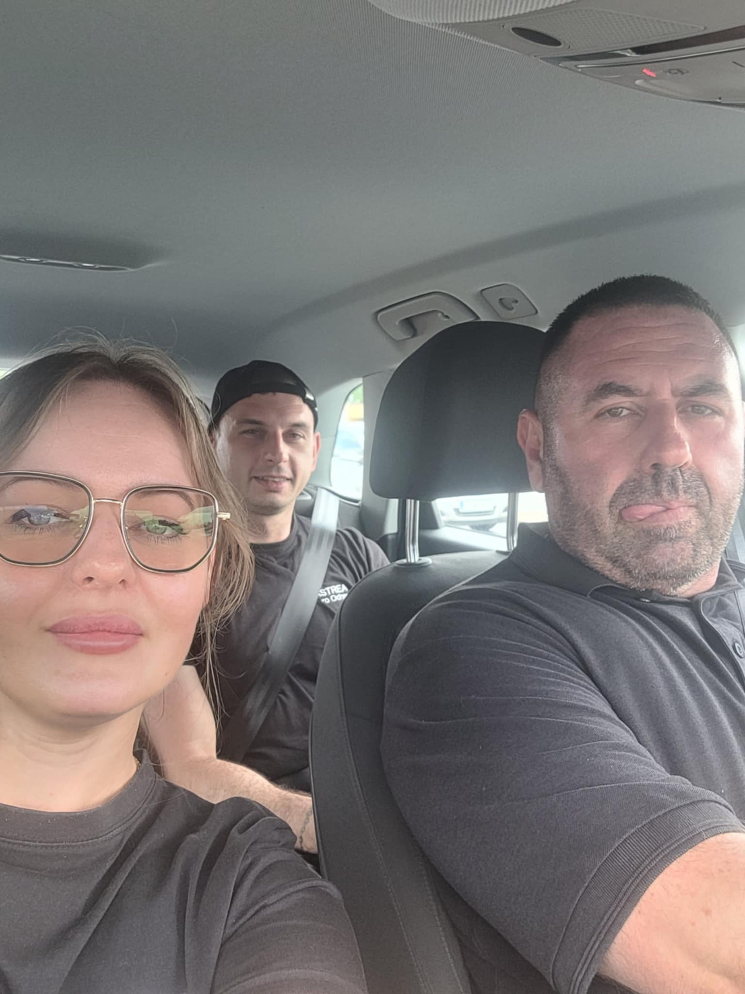 Trzech ochroniarzy w czarnych ubraniach siedzi w samochodzie, prawdopodobnie w drodze na interwencję. Kierowca z brodą pokazuje język.