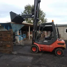 Czerwony wózek widłowy Linde z podniesioną łyżką stoi na placu obok drewnianych bali i niebieskiego podajnika, w tle zadaszony skład z workami.