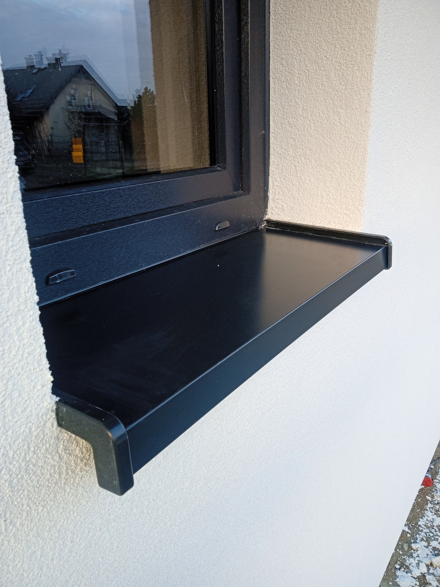 Zamontowany, czarny parapet zewnętrzny przy oknie na tle jasnej, chropowatej elewacji budynku. Widoczne odbicie domu w szybie okna.
