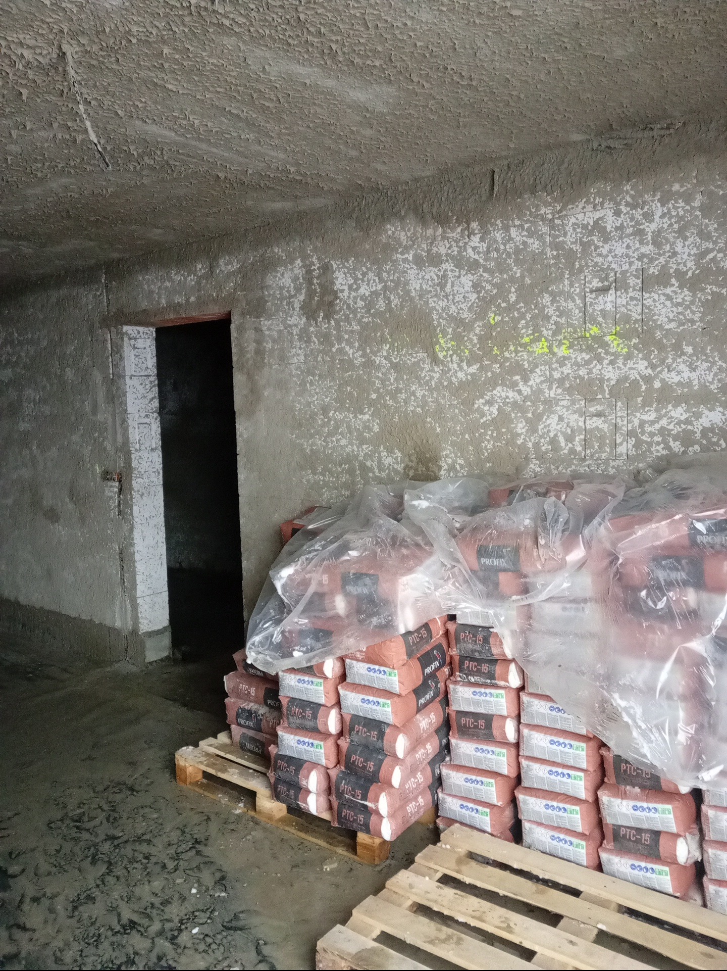 Stos palet z workami zaprawy PTC-15 w surowym, oświetlonym pomieszczeniu z tynkowanymi ścianami i sufitem. Widoczne ślady po łatach i żółte oznaczenia na ścianie.