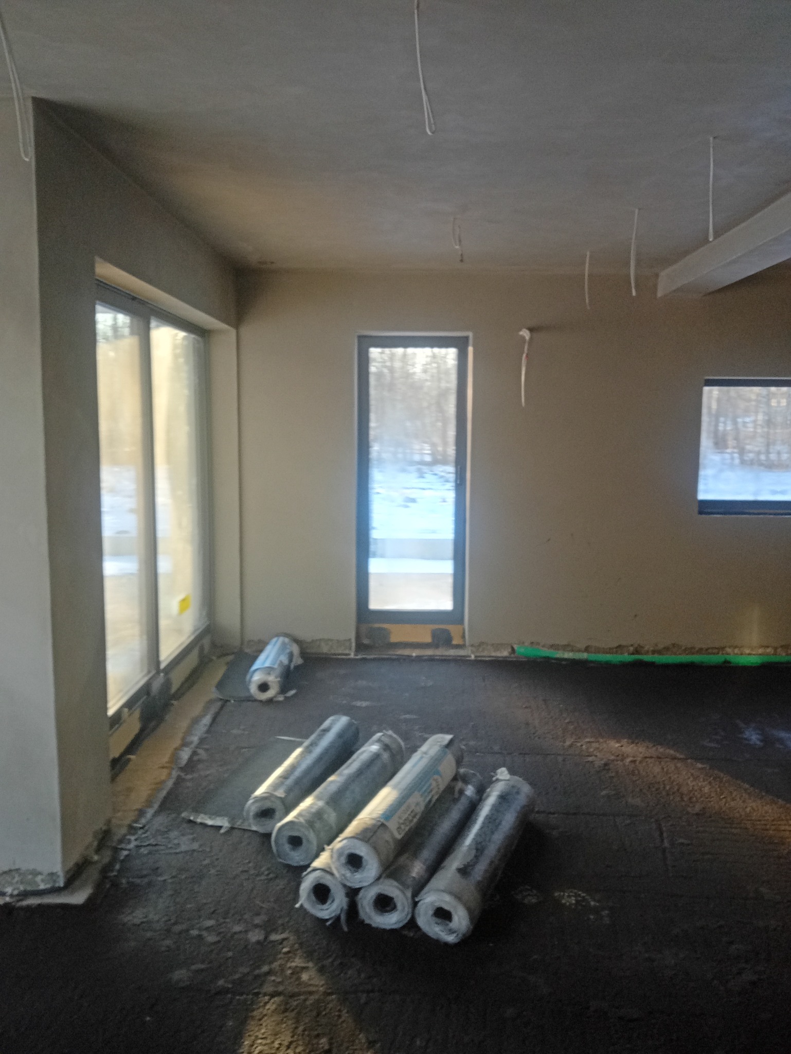 Przygotowanie do izolacji podłogi w surowym wnętrzu: rozłożona papa termozgrzewalna i zrolowane materiały izolacyjne. Widoczne okna i drzwi z widokiem na zimowy krajobraz.