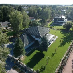 Nowoczesny dom jednorodzinny z ciemną dachówką, widok z lotu ptaka, zadbany trawnik, ogrodzenie z elementami betonowymi i nasadzeniami, w tle zabudowa mieszkalna i drzewa.