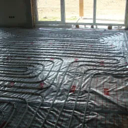 Ułożone rury ogrzewania podłogowego na folii z czerwoną siatką w surowym wnętrzu. Widoczne okno i betonowe ściany w stanie deweloperskim.