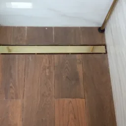 Szczegół odpływu liniowego w podłodze kabiny prysznicowej, wykończonej drewnopodobnymi płytkami, z widocznymi elementami szklanej kabiny i białymi płytkami ściennymi.