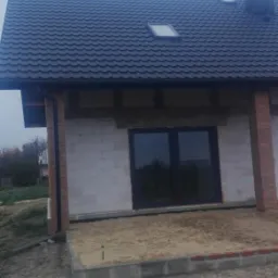 Dom w stanie surowym zamkniętym z ciemną dachówką, widoczne okno dachowe, komin i fragment elewacji z cegły klinkierowej, teren wokół domu niezagospodarowany.