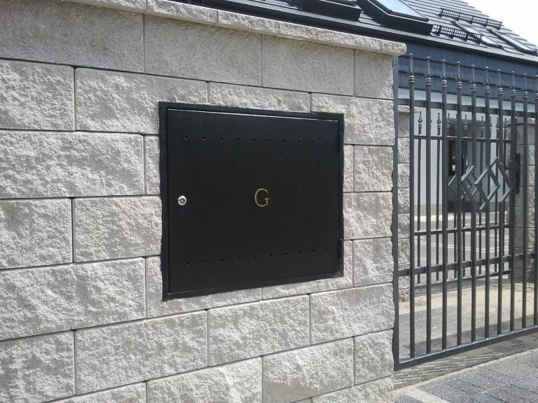 Czarna skrzynka z literą 'G' na kamiennym murze obok metalowej bramy wjazdowej.