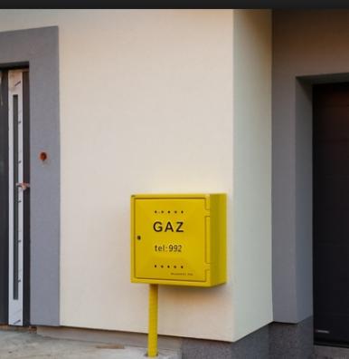 Żółta skrzynka gazowa z napisem GAZ i numerem telefonu 992, umieszczona na elewacji budynku przy wejściu.