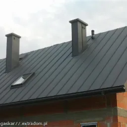 Szary dach z blachy na domu w trakcie budowy, widoczne kominy i okno dachowe.