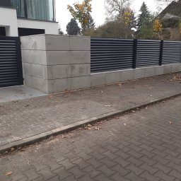 ABG MATERIAŁY Bartosz Lewandowski - Nowoczesne ogrodzenie z poziomych, czarnych paneli zamontowanych na betonowych słupkach i podmurówce, z bramką wejściową i kostką brukową na pierwszym planie, jesienne liście na chodniku.