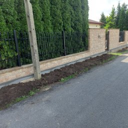 ABG MATERIAŁY Bartosz Lewandowski - Fragment ogrodzenia z elementami kutymi i betonowymi, obok świeżo nasypana ziemia, w tle wysokie tuje.