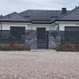 Nowoczesny dom jednorodzinny z grafitową, poziomą, metalową zabudową ogrodzeniową na kamiennej podmurówce, z bramką wejściową i nasadzeniami z czerwonych krzewów.