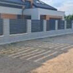 ABG MATERIAŁY Bartosz Lewandowski - Nowoczesne ogrodzenie z poziomych, szarych paneli między betonowymi słupkami, widok z zewnątrz posesji, cień rzucany przez panele na piaszczyste podłoże.