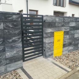 ABG MATERIAŁY Bartosz Lewandowski - Nowoczesne ogrodzenie z szarych, betonowych bloczków z poziomą, metalową bramką; żółta skrzynka gazowa zamontowana na ogrodzeniu; ścieżka z jasnej kostki brukowej z ciemnymi obrzeżami.
