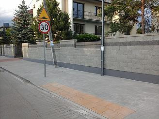 Nowoczesne ogrodzenie z szarej kostki betonowej z elementami dekoracyjnymi w postaci ciemniejszych pasów, widok z poziomu chodnika, w tle dom jednorodzinny z balkonem i zielone drzewa iglaste, po...