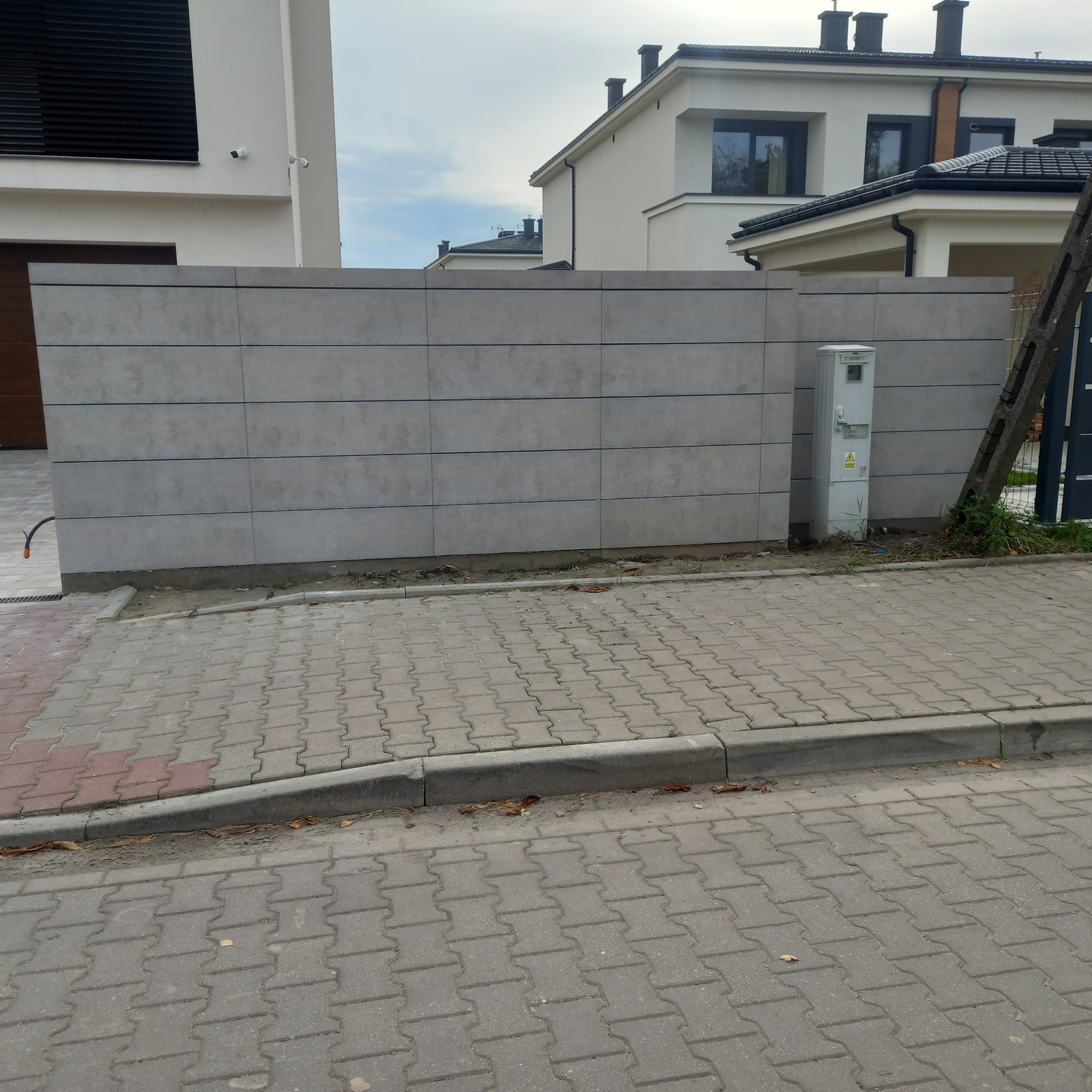Nowoczesne ogrodzenie betonowe z poziomymi panelami w odcieniach szarości, widoczne z poziomu chodnika z kostki brukowej, z budynkami w tle i skrzynką elektryczną przy słupie.