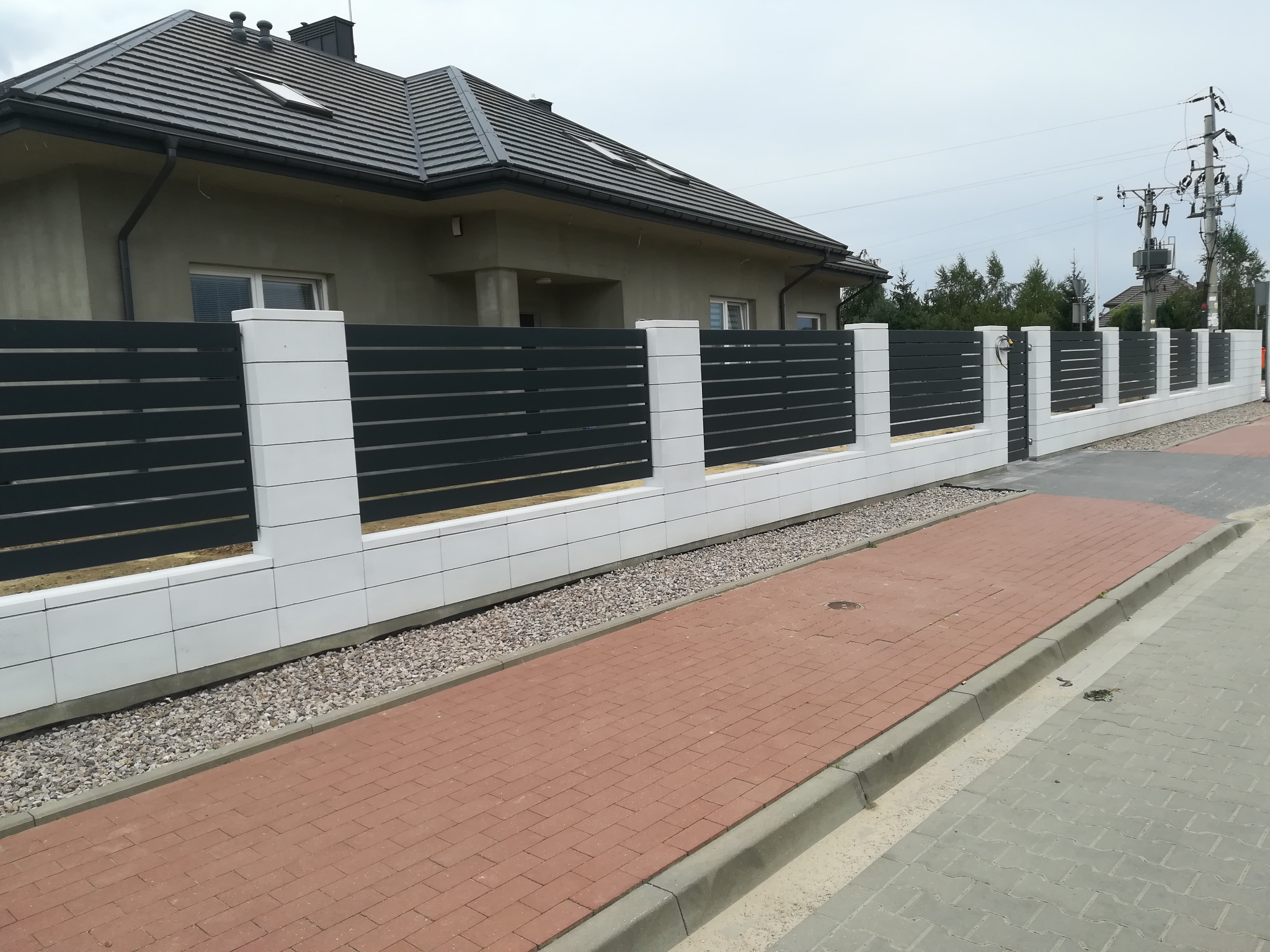 Nowoczesne ogrodzenie posesji z poziomych, grafitowych paneli między białymi, modułowymi słupkami, widok z chodnika wyłożonego czerwoną kostką brukową.