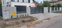 Nowoczesne ogrodzenie panelowe z poziomych profili w kolorze antracytowym, osadzone na betonowych słupkach, z kostką brukową na podjeździe i młodymi roślinami u podstawy.