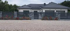 Nowoczesny dom jednorodzinny z grafitową, poziomą, metalową zabudową ogrodzeniową na kamiennej podmurówce, z bramką wejściową i nasadzeniami z czerwonych krzewów.