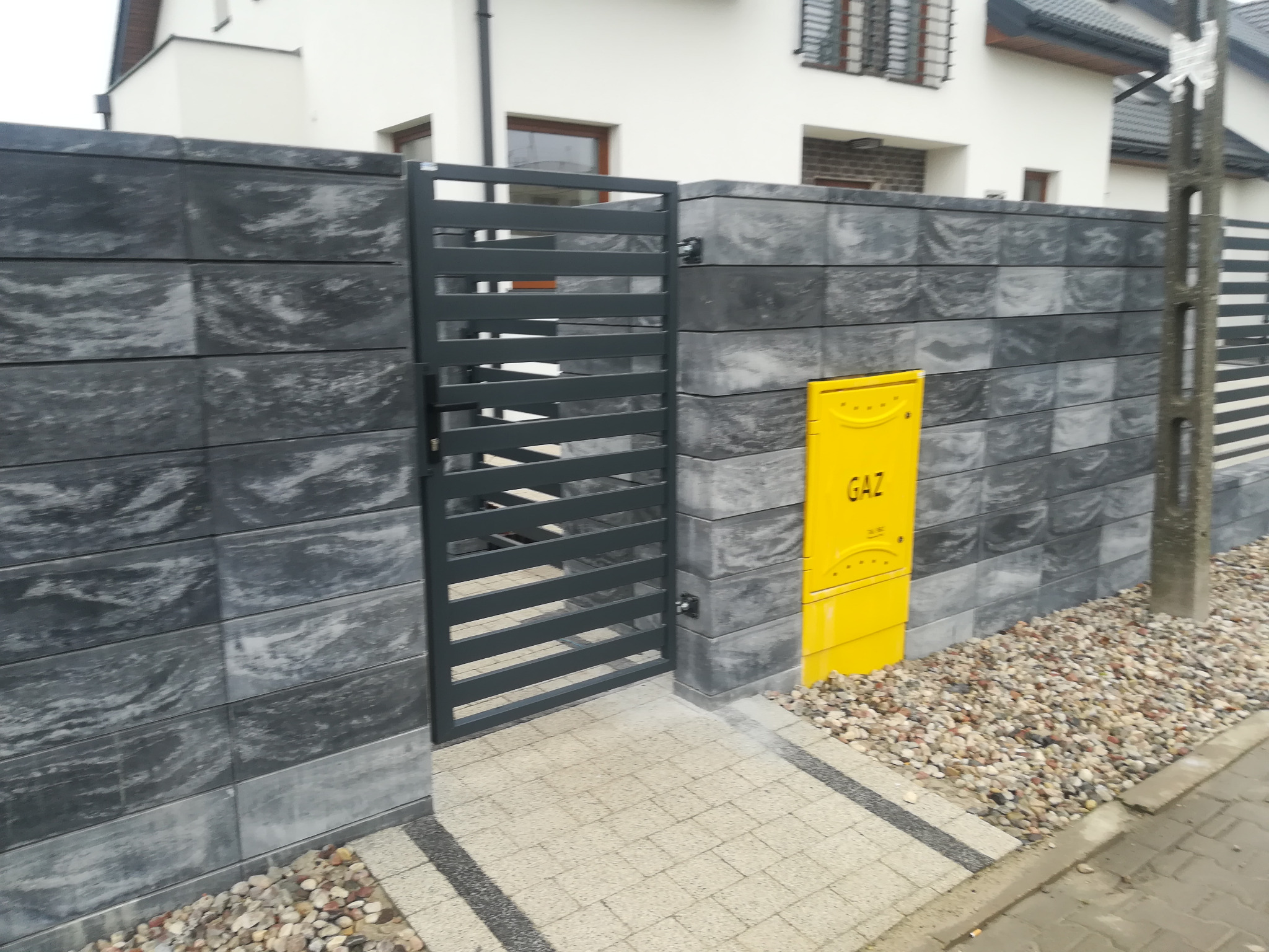 Nowoczesne ogrodzenie z szarych, betonowych bloczków z poziomą, metalową bramką; żółta skrzynka gazowa zamontowana na ogrodzeniu; ścieżka z jasnej kostki brukowej z ciemnymi obrzeżami.
