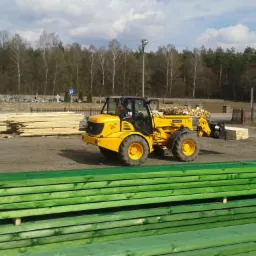 Żółty wózek widłowy JCB z ładunkiem drewna na placu, w tle stosy tarcicy impregnowanej na zielono, niezabezpieczone drewno, las i cmentarz.