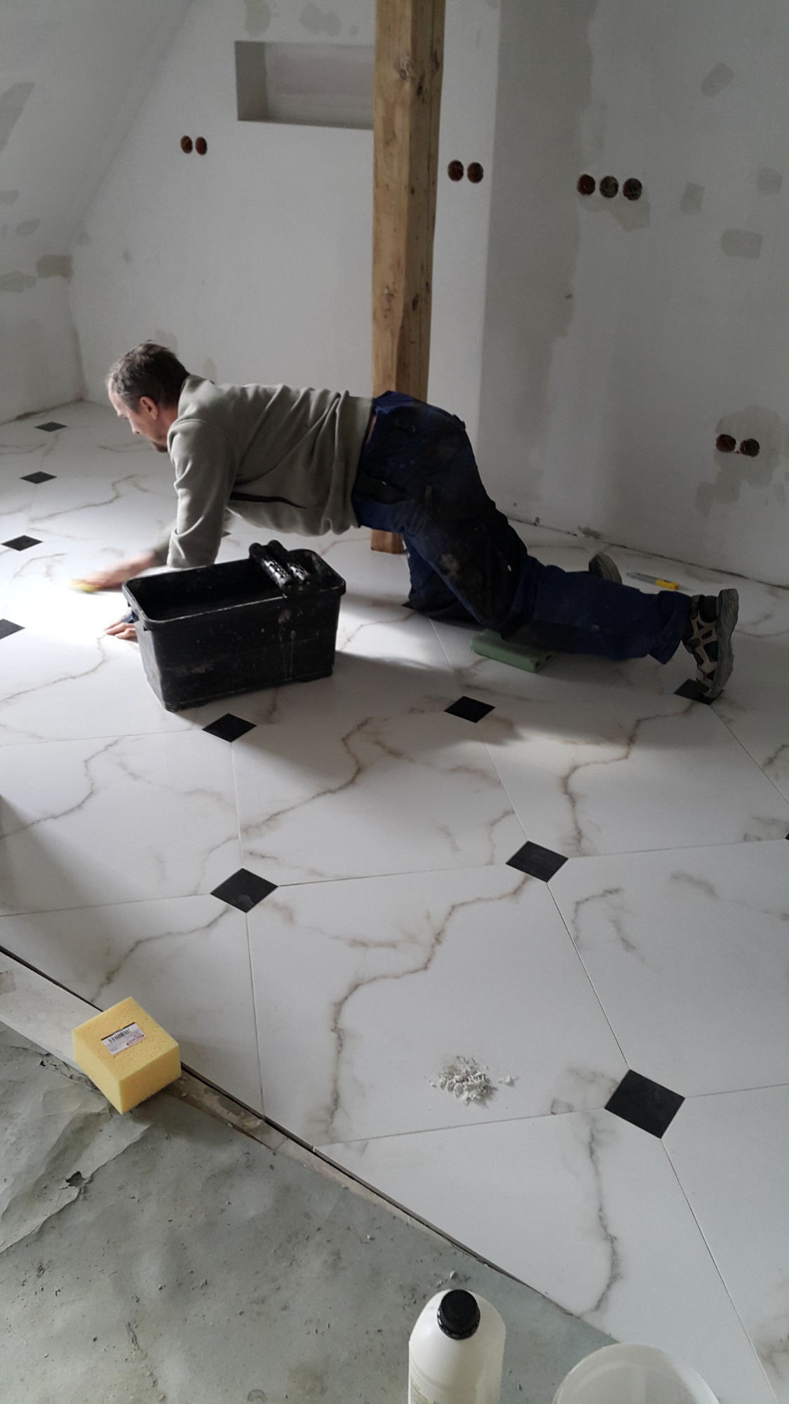 Mężczyzna w trakcie fugowania białych płytek podłogowych z czarnymi wstawkami w pomieszczeniu ze skośnym sufitem i drewnianą belką konstrukcyjną.