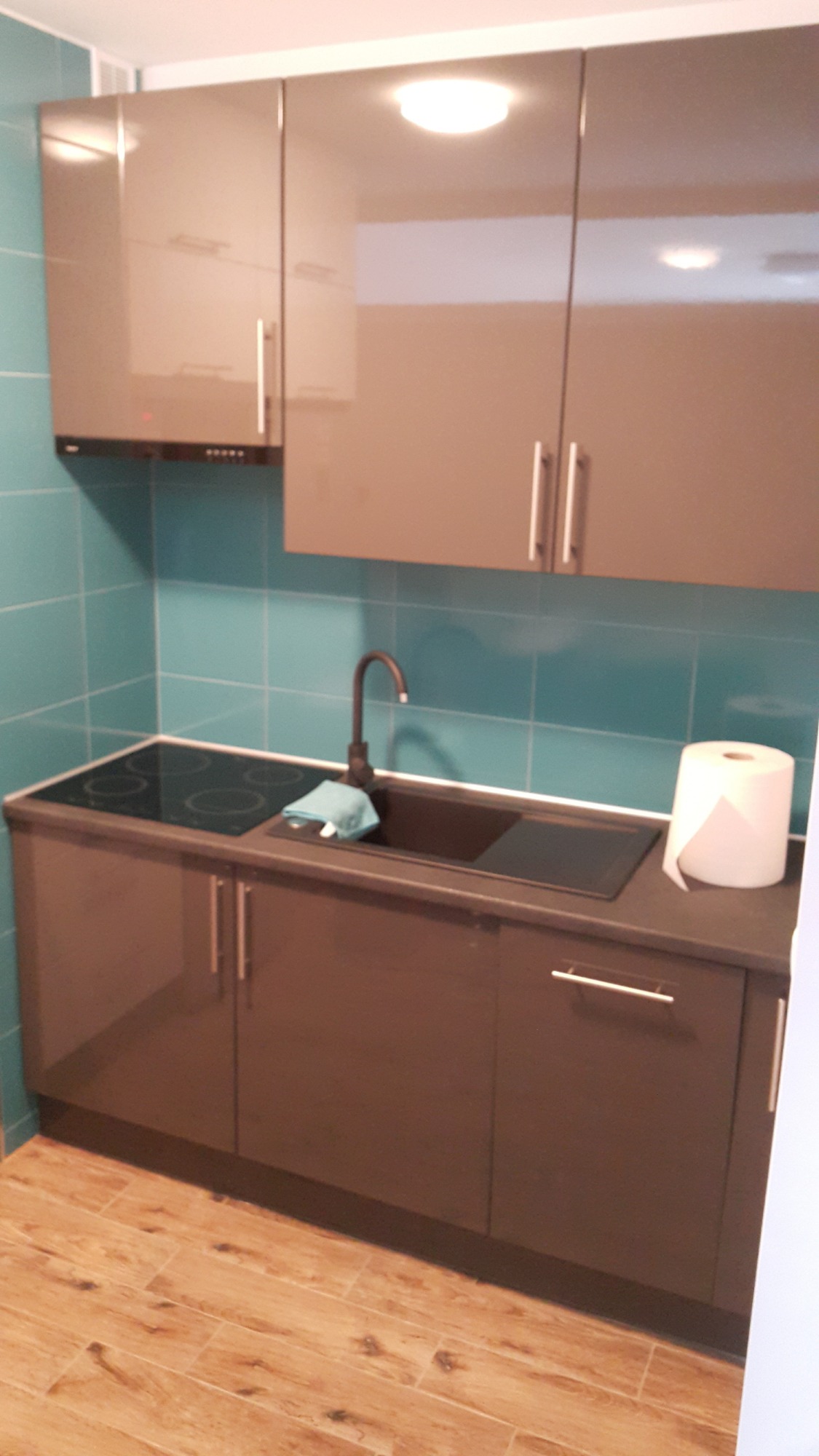 Nowoczesna kuchnia w Krakowie po remoncie: błyszczące, grafitowe szafki górne z ukrytym okapem, turkusowe płytki na ścianie, zintegrowana płyta grzewcza i zlewozmywak z czarną armaturą,...