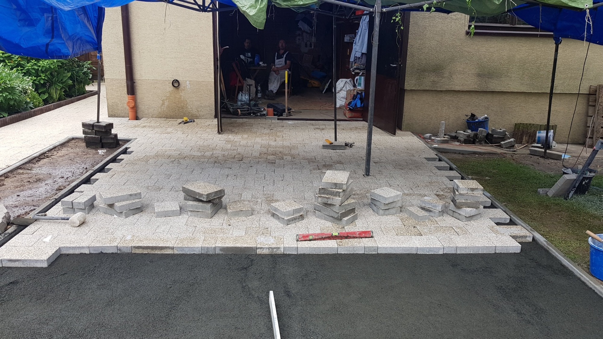 Układanie kostki brukowej na przygotowanym podłożu, widoczny fragment ułożonej nawierzchni oraz stosy kostki, w tle garaż i pracownicy w trakcie przerwy.
