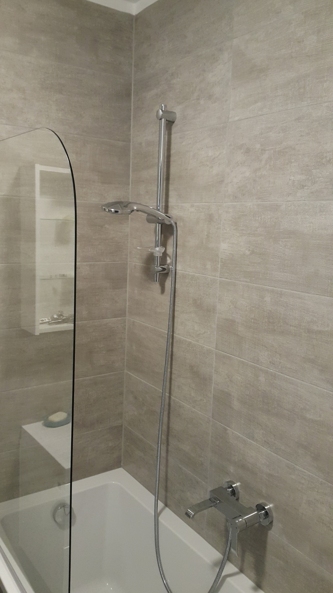 Nowoczesna kabina prysznicowa z beżowymi płytkami imitującymi drewno, chromowana bateria i deszczownica, widoczna półka z mydłem.