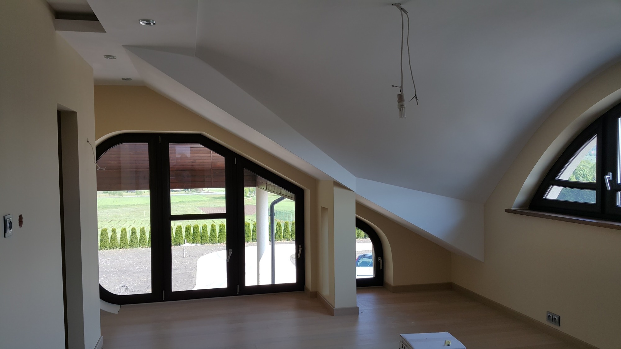 Poddasze z oknami o nietypowym kształcie po malowaniu ścian na jasny beż, widoczne niedokończone oświetlenie sufitowe i skrzynka na podłodze.