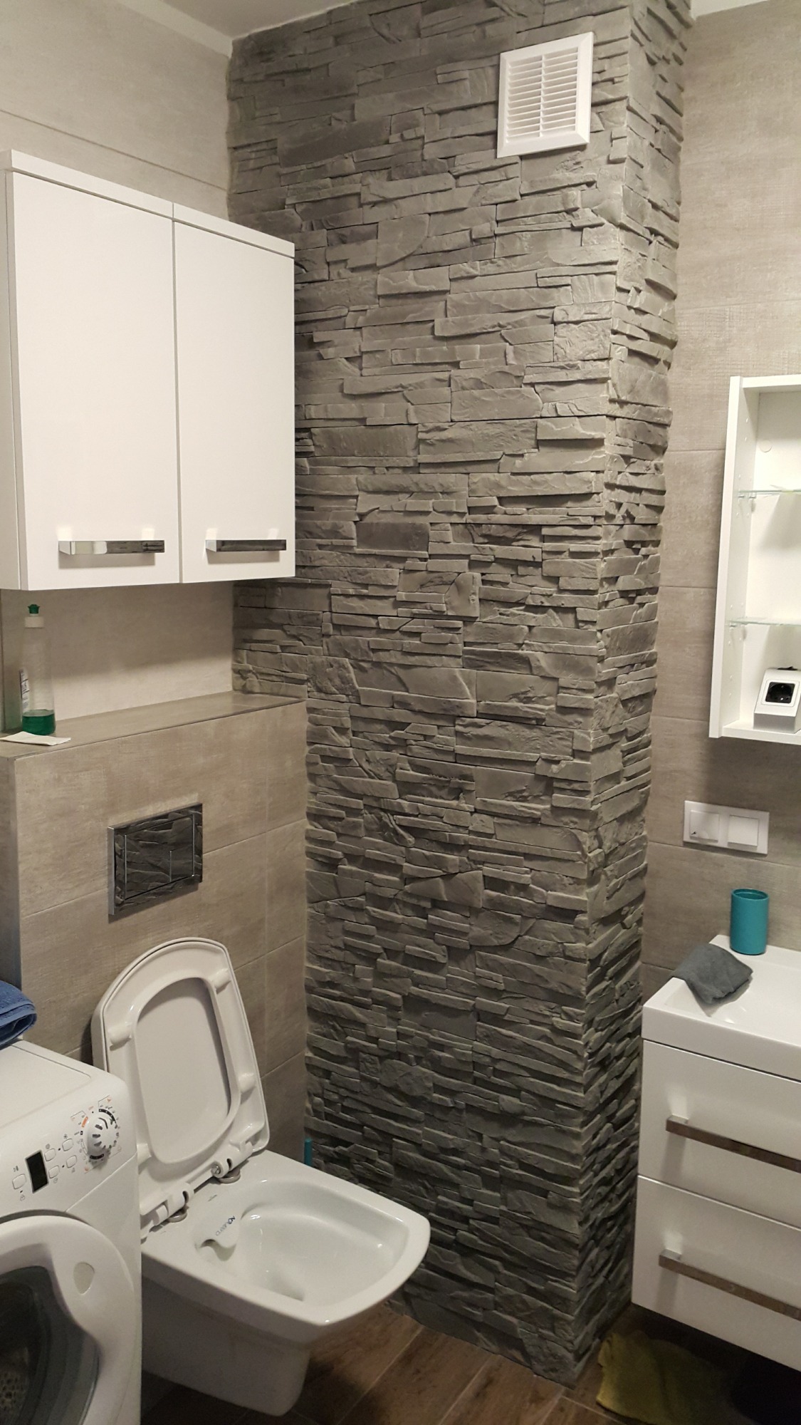 Wykończona łazienka z toaletą, pralką i umywalką, akcentowana kolumną z szarej, dekoracyjnej okładziny kamiennej na ścianie, oświetlona ciepłym światłem.