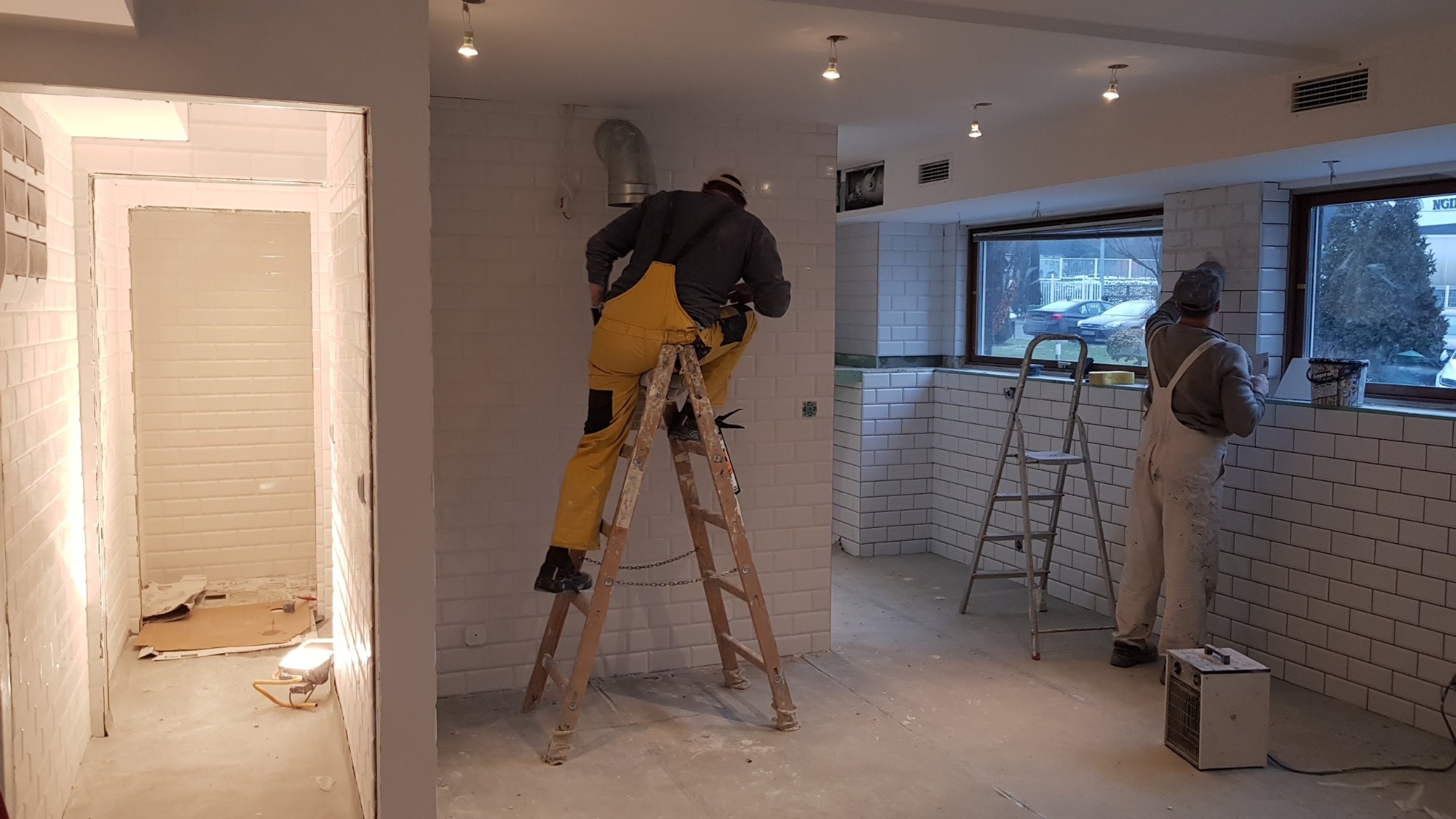 Dwóch pracowników podczas wykańczania wnętrza z białymi, prostokątnymi płytkami. Jeden na drewnianej drabinie w żółtym kombinezonie, drugi w białym kombinezonie przy oknie. Widoczny fragment...