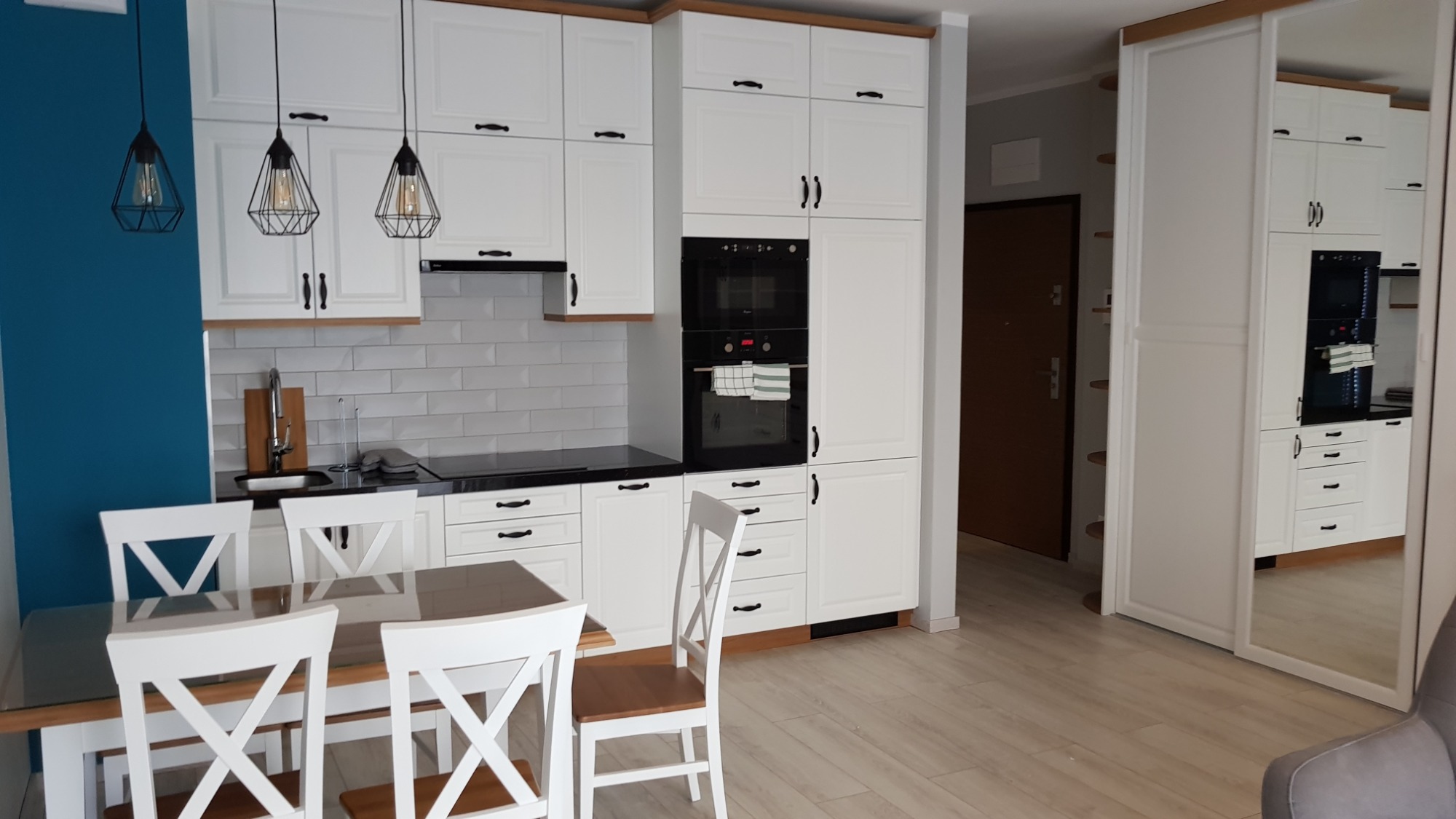 Jasna kuchnia z białymi meblami w stylu skandynawskim, czarnym blatem i trzema wiszącymi lampami o geometrycznych kloszach nad drewnianym stołem z białymi krzesłami.