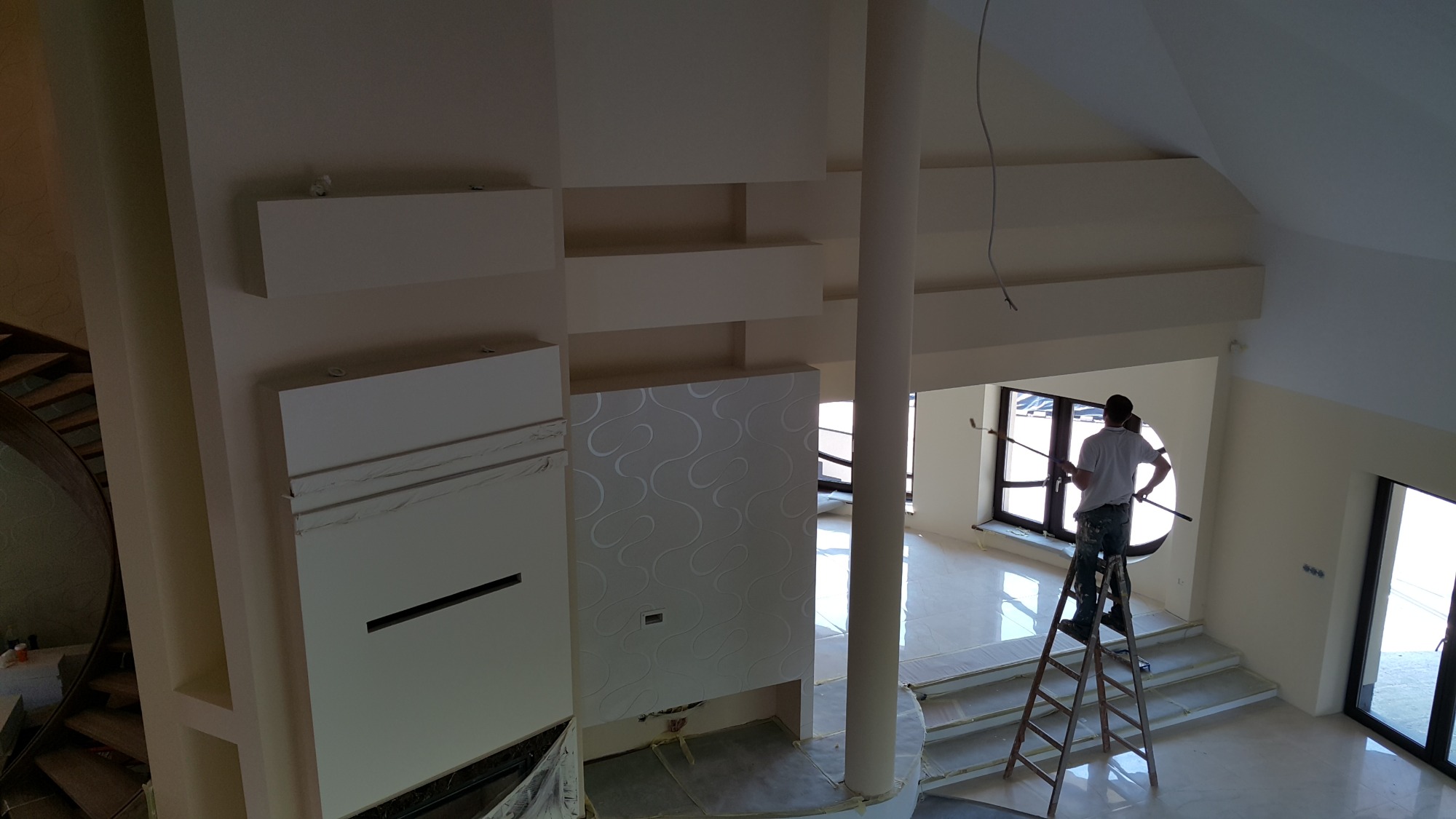 Mężczyzna na drabinie maluje ścianę w jasnym, nowoczesnym wnętrzu z okrągłym oknem i dekoracyjną ścianą z wzorem, widok z góry.