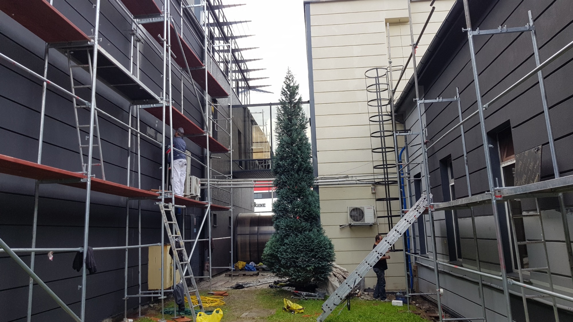 Prace na rusztowaniu przy elewacji budynku z ciemnymi panelami, widoczny pracownik w białych spodniach i drabina oparta o ścianę, na trawniku żółte worki i przewody.